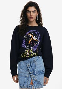 Sudadera negra con un diseño de cara abstracta y colorida. Combinada con una falda de mezclilla de bolsillo múltiple en un tono azul claro con estilo envolvente.
