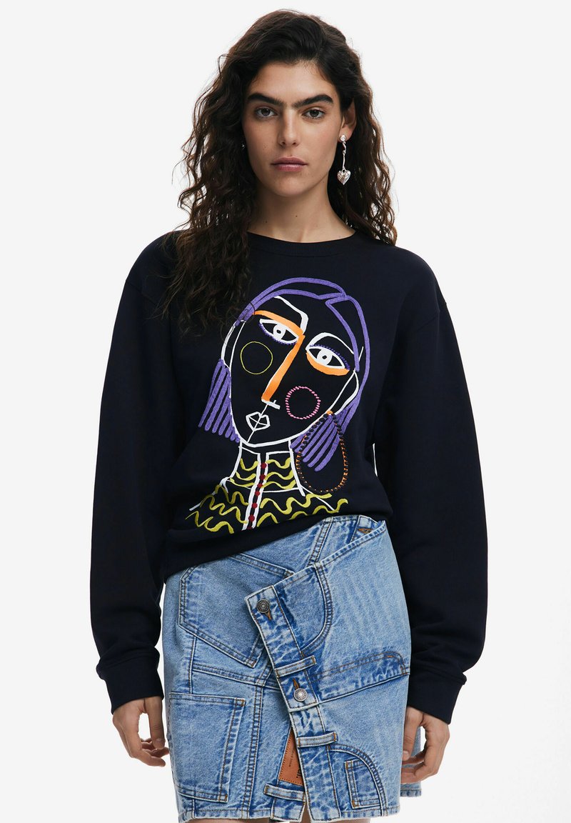 Sudadera negra con un diseño de cara abstracta y colorida. Combinada con una falda de mezclilla de bolsillo múltiple en un tono azul claro con estilo envolvente.