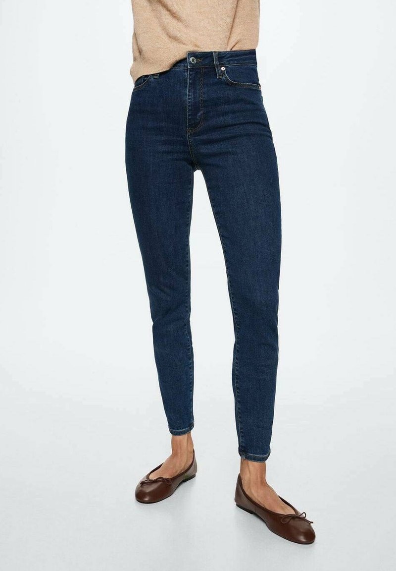 Personne portant un jean skinny taille haute bleu foncé et des ballerines marron, debout devant un fond clair uni.