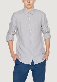 Camicia grigia chiaro con bottoni, a strisce verticali sottili; maniche lunghe arrotolate fino ai gomiti; il tessuto appare testurizzato e casual.