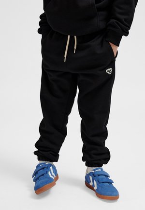 Zwarte katoenen joggers met elastische boorden en een verstelbare trekkoord, gecombineerd met blauwe klittenbandschoenen met witte accenten.