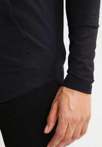 Fekete, hosszú ujjú felső, puha anyagból készült, kerekített hem elülső résszel és texturált felülettel, a csuklónál testhez simuló kialakítással.