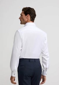 Camisa blanca de manga larga con cuello clásico, puños con botones, textura suave y diseño con yugo en la espalda, combinada con pantalones oscuros.