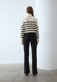 Kvinna med långt brunt hår står vänd mot en vit vägg, iklädd en krämfärgad polotröja med svarta ränder och svarta bootcut-jeans.