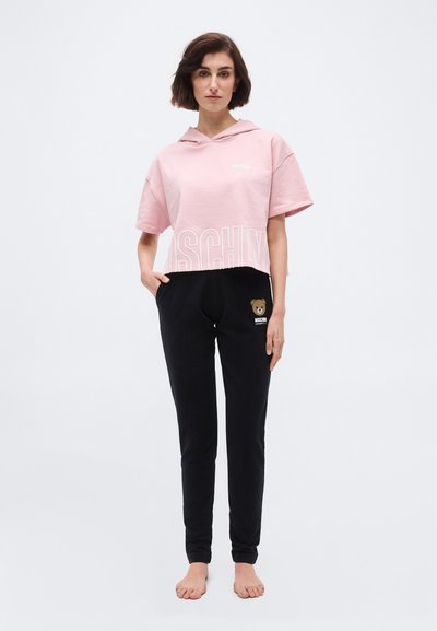 Γυναίκα που στέκεται ξυπόλητη φορώντας ένα ροζ κοντομάνικο φούτερ με κουκούλα και μαύρο παντελόνι με λογότυπο αρκουδάκι στο αριστερό μηρό.