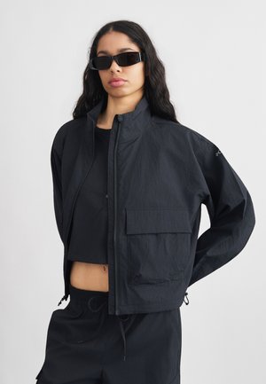 Kvinde iført sort oversized jakke, sort cropped top, sorte bukser og sorte rektangulære solbriller mod en ensartet hvid baggrund.