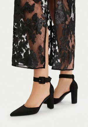 Piernas con zapatos de tacón grueso negro de punta puntiaguda y correas en el tobillo, con una falda de encaje negro translúcido con patrones florales y una abertura frontal.