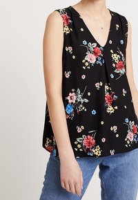 Frau in einem ärmellosen schwarzen Top mit roten, rosa, blauen und gelben Blumenmustern, kombiniert mit blauen Jeans und einer zarten Halskette.