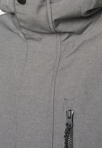Gros plan d'une veste en tissu gris montrant une fermeture éclair avec tirette et une partie du col montant avec un bouton-pression noir.