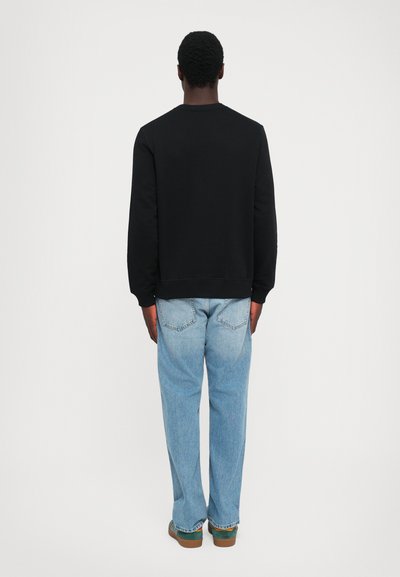 Man standing facing away wearing a black long-sleeve shirt, light blue jeans, and green sneakers against a plain white background.