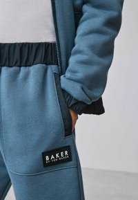 Blauwe textuur zip-up jack met elastische manchetten, gecombineerd met bijpassende joggers met een zwart merklabel. Zacht, casual materiaal.