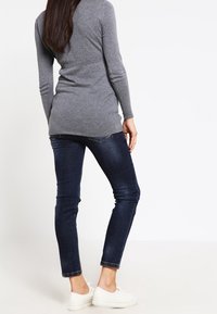 Grauer Langarmshirt, kombiniert mit dunkelblauen Skinny-Jeans und weißen Sneakers. Das Shirt hat einen figurbetonten Schnitt und einen leicht längeren Rücken.