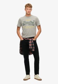 Camiseta gris de algodón con texto "SuperDry", jeans negros y camisa de franela a cuadros atada a la cintura. Zapatillas con cuerpo color crema y suela marrón.