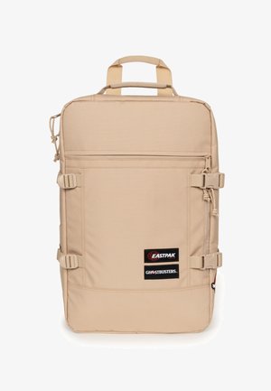 Zaino rettangolare beige con due toppe frontali che riportano la scritta "Eastpak" e "Ghostbusters", fibbie laterali regolabili e manici superiori.