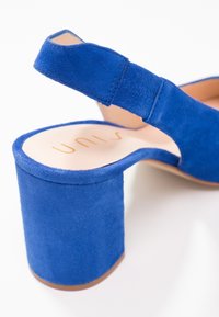 Chaussure à talons hauts en suède bleu avec un talon épais et une bride arrière. La semelle intérieure arbore un logo en lettres dorées. Surface lisse et texturée.