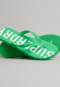 Groene rubberen slippers met een gestructureerde zool en dikke banden, met in grote witte letters "SUPERDRY" erop gedrukt.