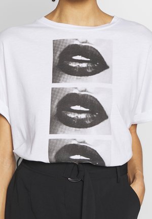 Wit T-shirt met drie zwart-wit close-up prints van lippen verticaal gestapeld, gedragen met een zwarte broek en riem.