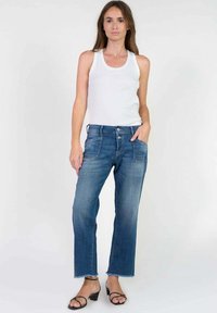 Un jean droit bleu clair présente un délavage, cinq poches et un ourlet brut. Associé à un débardeur blanc et des sandales noires.