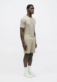 Bēša kokvilnas t-krekls ar izcirstu logotipu, komplektā ar atbilstošiem šortiem. Modelis valkā baltas zeķes un bēšas sporta kurpes. Vienkāršs, sportisks dizains.