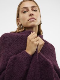 Lielgabarīta purpursarkans adīts džemperis ar ribotu tekstūru, augstu apkakli un apjomīgām piedurknēm, ko tur pie apkakles cilvēks.