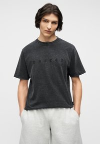 Νέος άνδρας με μακριά σκούρα μαλλιά μέχρι τους ώμους, φορώντας ξεθωριασμένο μαύρο μπλουζάκι με το κείμενο "WASTED" και ανοιχτόχρωμα γκρι σορτς, με τα χέρια στις τσέπες.