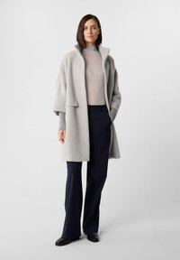 Donna che indossa un cappotto grigio chiaro in peluche sopra un dolcevita grigio, pantaloni a gamba larga blu navy e scarpe nere, in piedi su uno sfondo grigio uniforme.