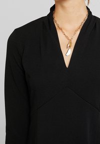 Mustaan tekstiilivaatteen, jossa on V-pääntie ja teksturoitu pinta. Korostaa kultasävyinen ketjun kaulakoru, jossa on kaksi koristeellista riipusta.