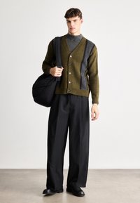 Πράσινος και γκρι ριγέ πλεκτός ζακέτα με κουμπιά, φορεμένος πάνω από ένα λαιμόκομμα. Χαλαρές μαύρες παν trousers και μια άνετη μαύρη τσάντα. Λείο υλικό και μοντέρνος σχεδιασμός.