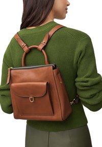 Sac à dos en cuir marron avec une forme structurée. Il présente une poche avant avec un rabat, des accents en métal doré et des bretelles réglables.