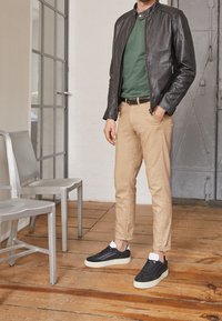 Černá kožená bunda, zelená košile, khaki chinos a černé tenisky s bílými akcenty, to vše na neutrálním vnitřním pozadí.