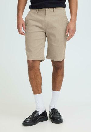 Uomo che indossa pantaloncini beige testurizzati, calzini bianchi alla caviglia e mocassini di pelle neri, in piedi contro uno sfondo chiaro e uniforme.