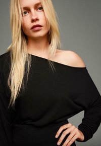 Čierny top s odhalenými ramenami a dlhými rukávmi, vyrobený z jemnej látky a uvoľneného strihu. Modelka má blond vlasy a neutrálne pozadie.