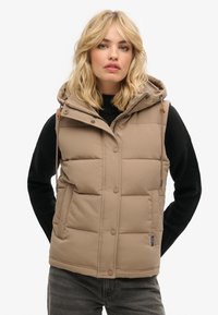 Beige Daunenweste mit Kapuze, gestepptem Design, vorderem Druckknopfverschluss und verstellbaren Kordeln. Über einem schwarzen Pullover getragen.