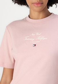 Roza majica kratkih rukava od pamuka s bijelim kurzivnim natpisom "New York Tommy Hilfiger" i malom oznakom loga.