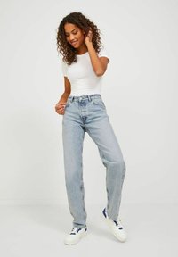 Šviesiai mėlynos aukšto liemens džinsai su tiesiu kirpimu, pagaminti iš džinsinio audinio, derinami su aptempta balta marškinėliais ir baltais sportiniais bateliais su mėlynomis detalėmis.