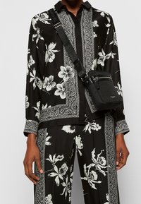 Zwart bloempatronen overhemd en broek met witte bloemen, voorzien van paisley-accenten en een relaxte pasvorm, gecombineerd met een kleine zwarte crossbody tas.