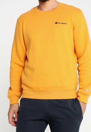 Mężczyzna noszący musztardową bluzę Champion z logo na piersi i rękawie, w połączeniu z ciemnogranatowymi spodniami.