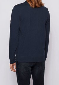 Sudadera de cuello redondo en color navy con mangas largas, puños y dobladillo acanalados. Presenta un parche de logo negro en el hombro izquierdo y una textura de algodón.