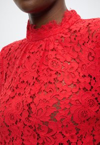 Czerwona koronkowa bluzka z wysokim kołnierzem i kwiatowymi wzorami. Tekstura jest misterna, z warstwowymi zdobieniami i falistymi krawędziami.