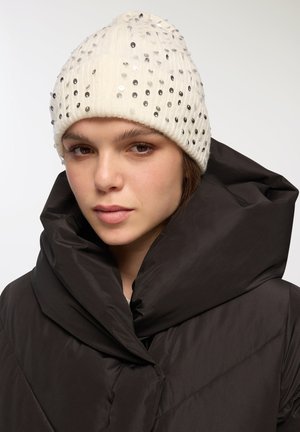 Berretto di maglia crema con paillettes, caratterizzato da una texture a coste. Abbinato a un giubbotto scuro e imbottito con un ampio colletto ripiegato.