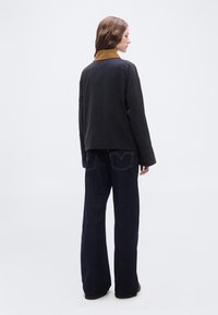 Γυναίκα με μακριά καστανά μαλλιά, πλαγιασμένη, που φοράει μαύρο μπουφάν με καφέ γιακά και σκουρόχρωμο τζιν με φαρδιά μπατζάκια, μπροστά σε απλό φόντο.