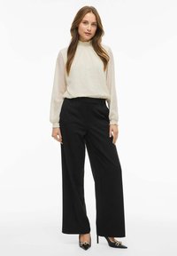 Cremfarvet bluse med teksturerede prikker, lange ærmer og flæse halsudskæring, parret med sorte brede bukser og sorte højhælede sko.