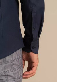 Mehe käsi puhkab kõrval, kannab tumesinist nupuga särkvarrast ja halli ruudulist pükste taustal beežil.