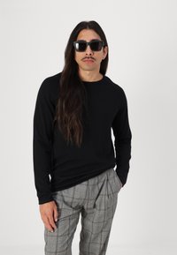 Cămașă neagră cu mânecă lungă și guler rotund, asortată cu pantaloni gri cu carouri și pliseuri; model purtând ochelari de soare negri.