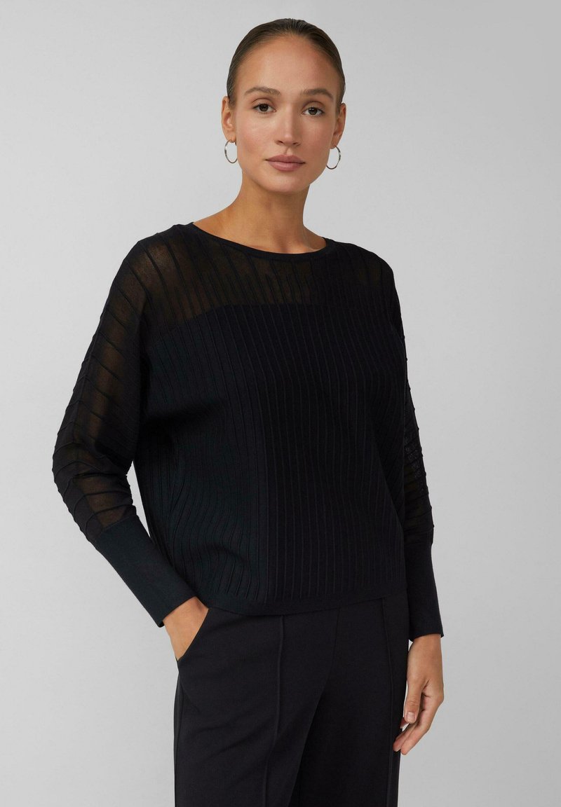 Maglione nero a coste con maniche lunghe trasparenti e scollo rotondo. Morbida texture con vestibilità comoda, dotato di tasche laterali.