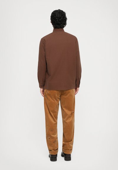 Homme avec des cheveux foncés et bouclés se tient de dos, portant une chemise à manches longues marron, un pantalon en velours côtelé moutarde avec des petites broderies florales et des chaussures noires.