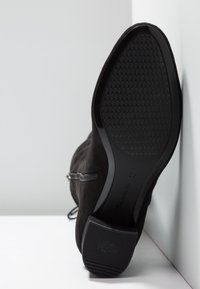 Bottine en suédine noire, légèrement inclinée contre un mur, montrant la semelle texturée et la fermeture éclair latérale, taille 37 visible sur la semelle.