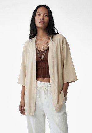 Kvinde med langt sort hår iført lagdelte halskæder, beige jakke med åben front, brun ribbet cropped top og hvide bukser med snor i taljen.