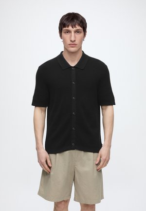 Ung mand iført en sort kortærmet polo med knapper og beige knælange shorts mod en hvid baggrund.