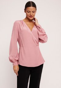 Różowa satynowa bluzka z długimi, bufkami rękawami i głębokim dekoltem w kształcie litery V. Posiada dopasowaną talię oraz guziki przy mankietach jako detal.
